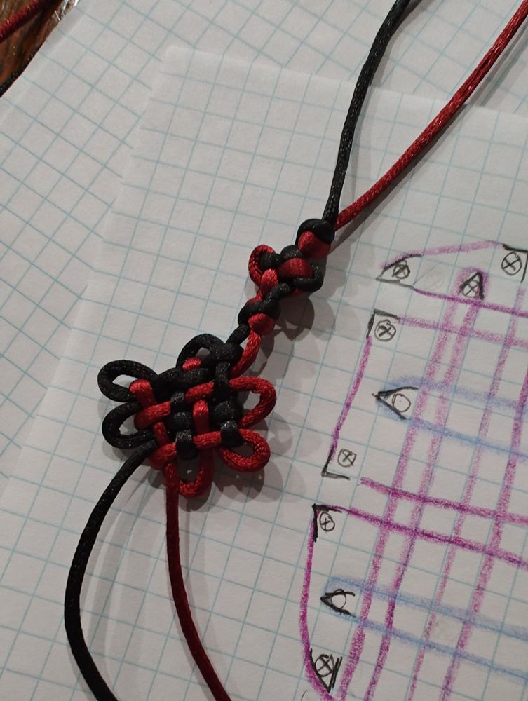 A red and black in-progress knotted bracelet sitting on graph paper, the corner of the paper shows a sketch I made to try to understand the chrysanthemum knot instructions.
