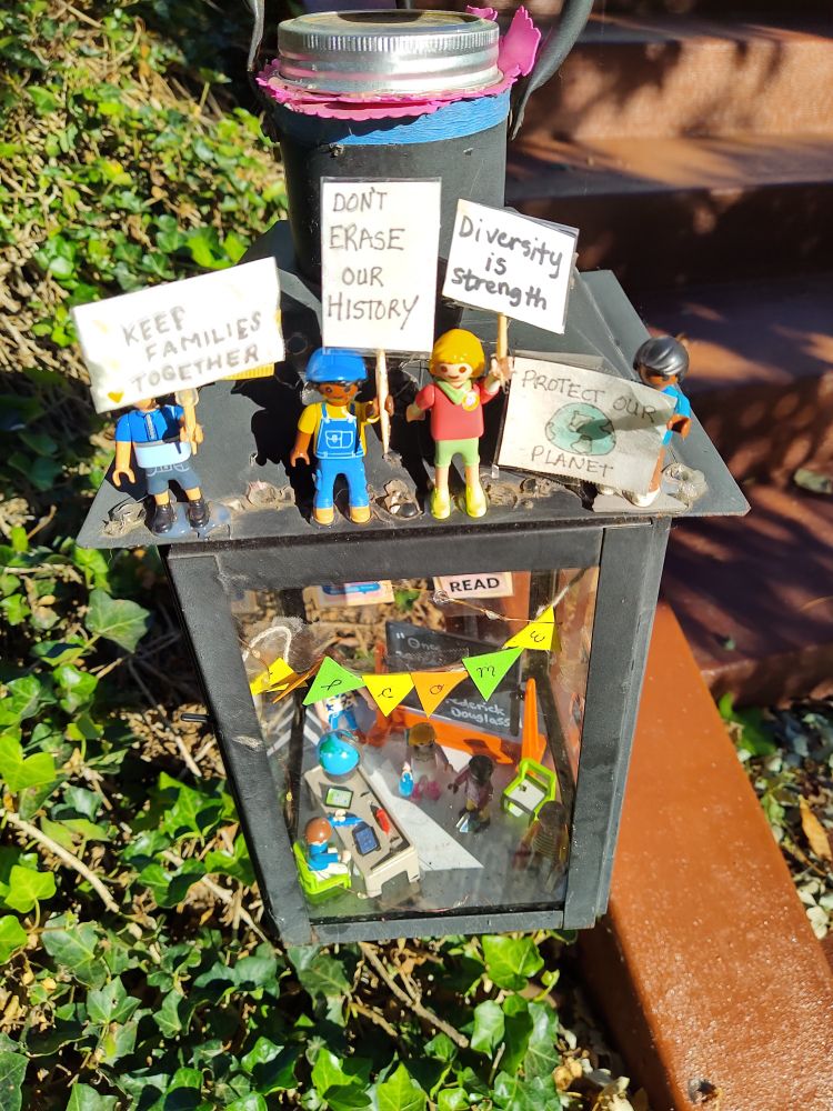 diorama on sidewalk with little figures protesting with signs like Keep Families Together, Don't Erase Our History, and Diversity is strength.