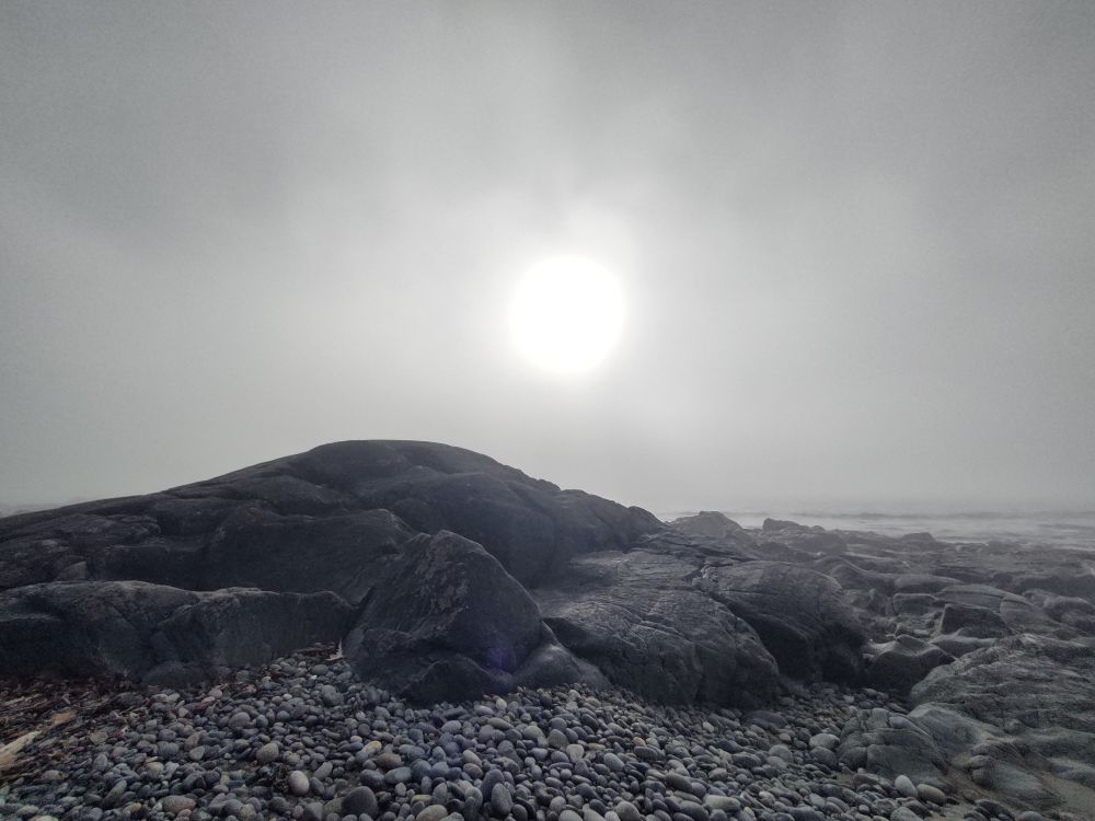 baleful sun through mist with rocks and gravel on beach below