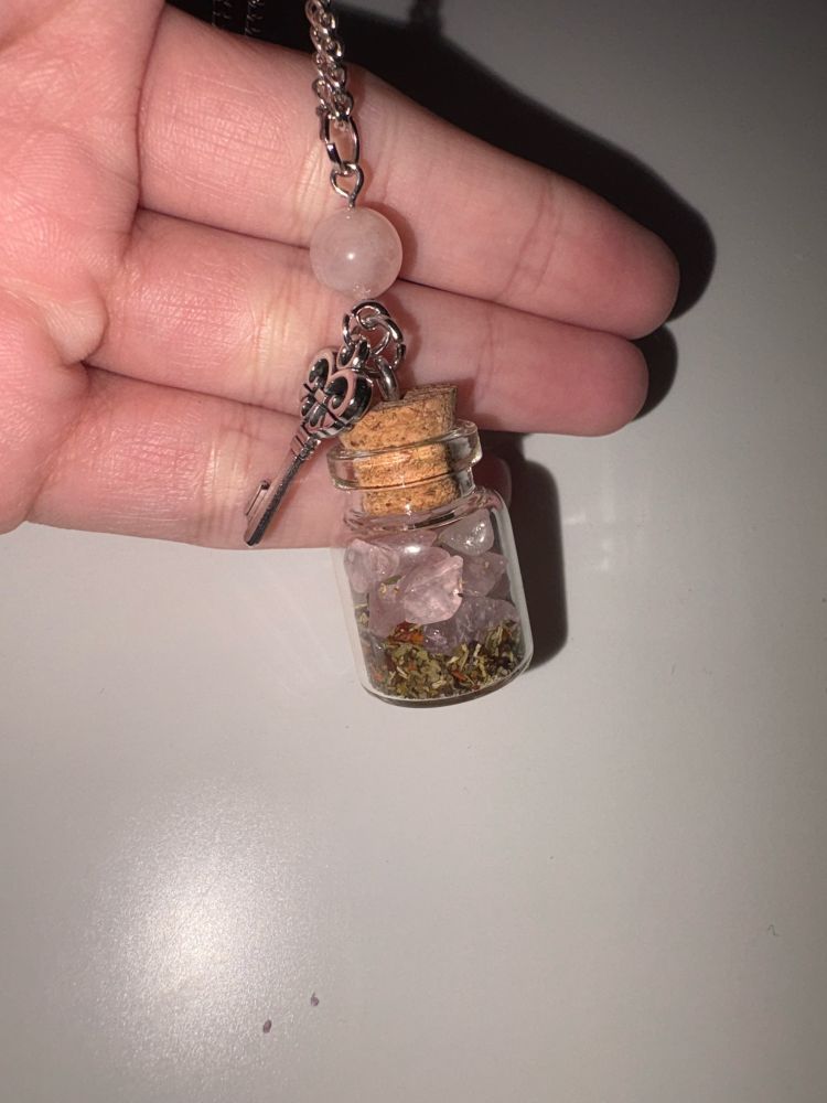 Tiny glass bottle pendant filled with herbs and pink crystals, closed with cork. Embellished with a silver, key shaped charm, and a crystal bead.