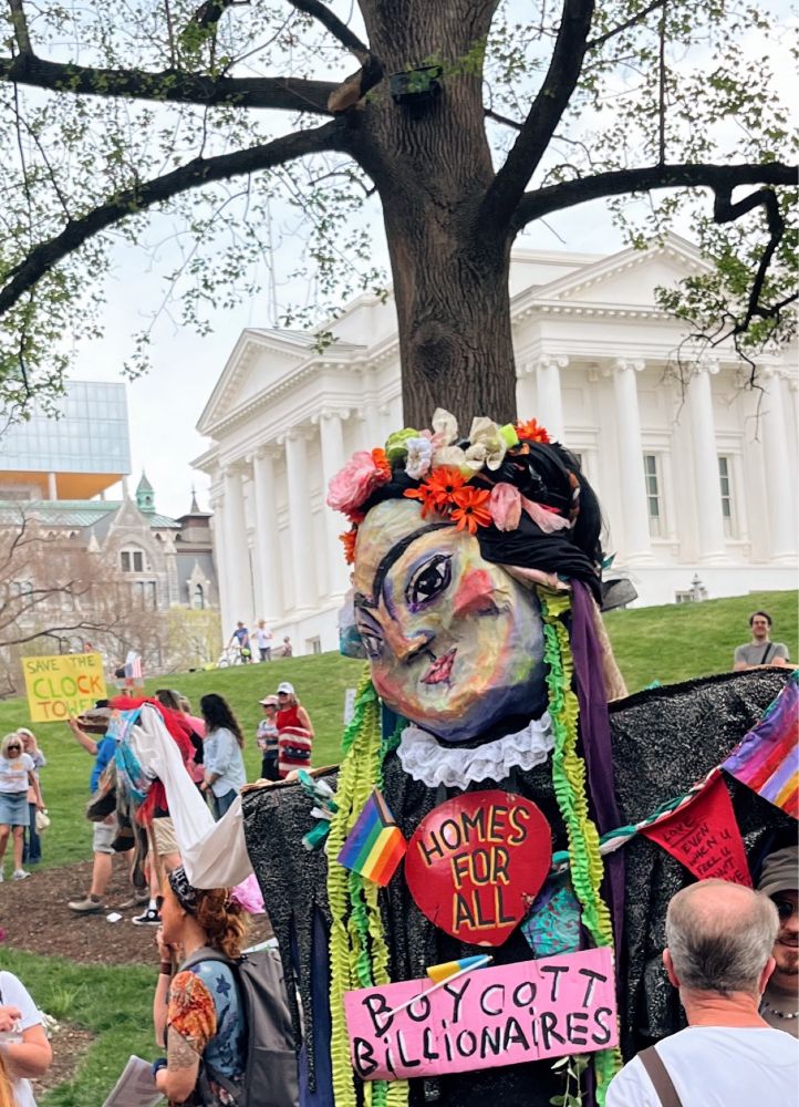 A Frida Kahlo puppet wearing signs that say Homes for All and Boycott Billionaires 
