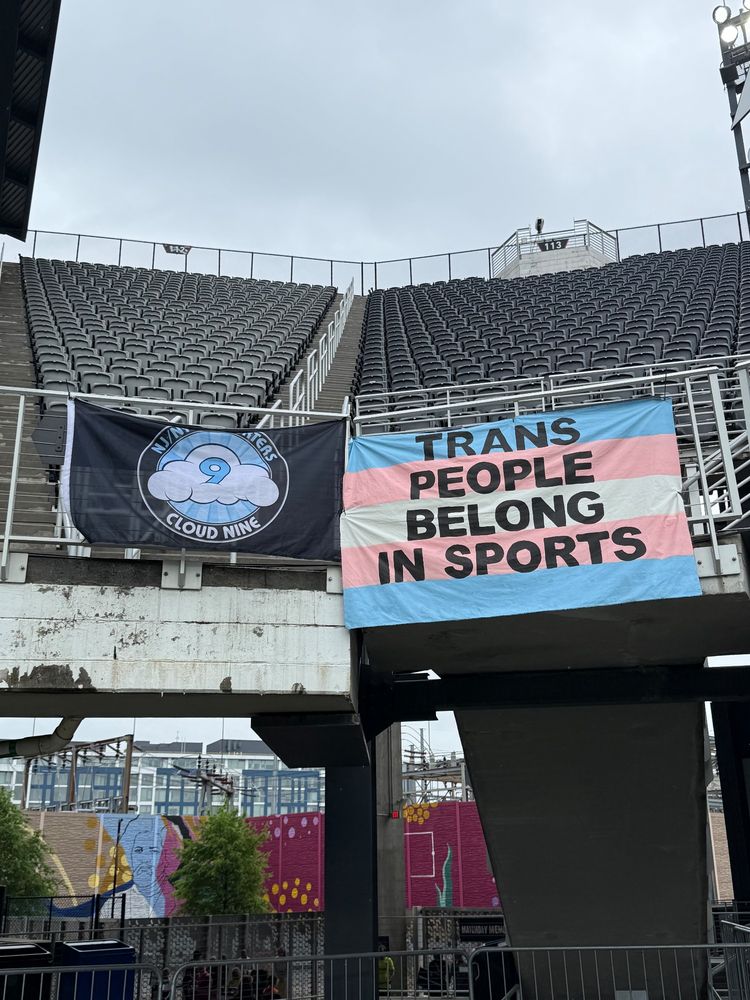 Photo of banners at Audi Field prior to the Gotham vs Spirit game. The banner on the right has trans pride colors with black text that reads Trans People Belong In Sports. To the left of the banner is a black flag with the Cloud 9 logo in the center.