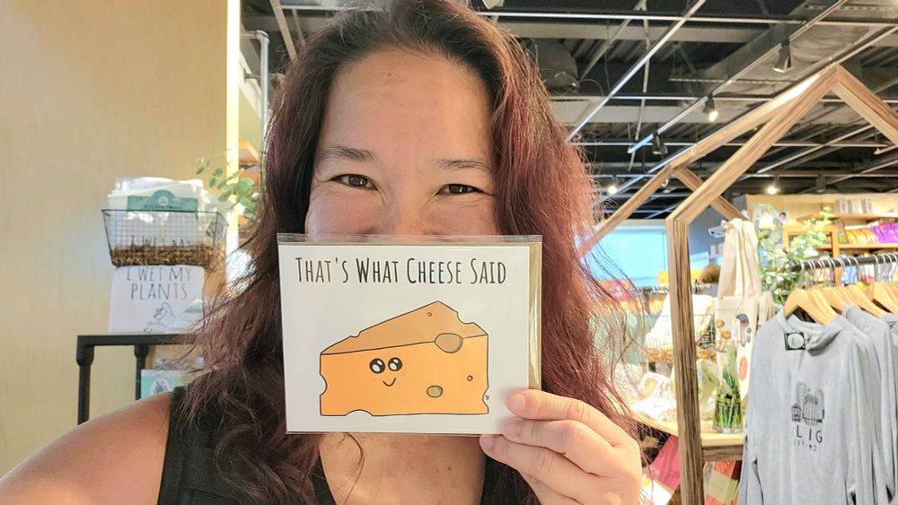 Selfie of me, a Chinese-American woman, holding up a card in front of my face that has a drawing of a wedge of cheese with a happy face and text that reads "that's what cheese said"