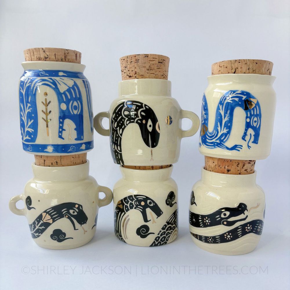 A collection of six white ceramic jars stacked on top of each other. They all feature unique, snake inspired designs. They were painted with blue or black underglazes and finished with gold luster details.