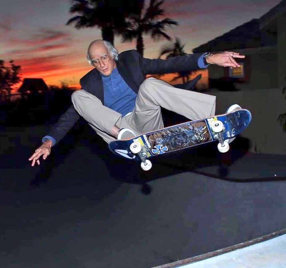 A picture of Tony Hawk in his Larry David costume skateboarding making a "Larry David"-esque facial expression. idk what the technical skate term is for what he's doing here in the picture, but it looks like he's up in the air defying gravity on his board, and you can see the bottom graphic on the board itself.