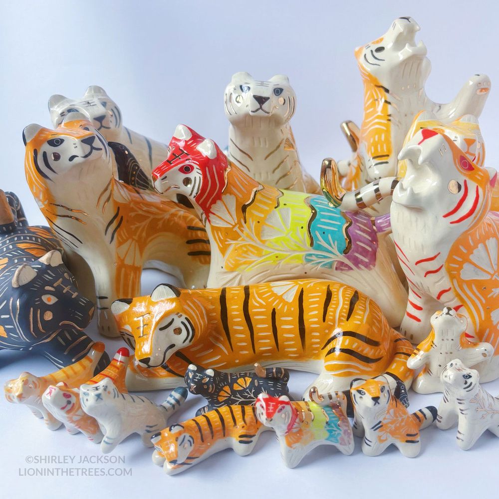 Family photo of 18 tigers—there are 9 large sized ceramic tigers and then 9 smaller mini-me's of the larger tigers. They feature a variety of designs and colours such as the traditional tiger colour, white, black, and a rainbow.