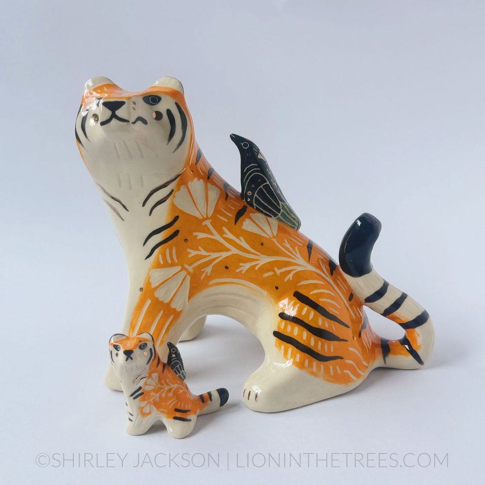 A large and mini me version of an orange and black ceramic tiger. They both have a magpie on their backs and they are both making the same "side-eye" face towards the magpie perched onto their backs.