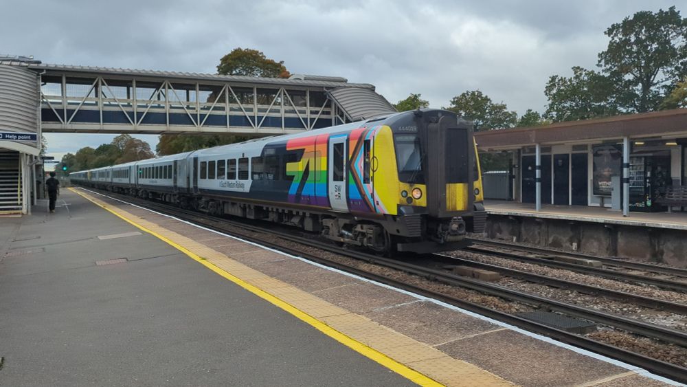 BR Class 444019; a five car South Western Railway 3rd rail powered EMU adorned with a pride flag passes West Byfleet Station on the South Western Mainline, UK. 