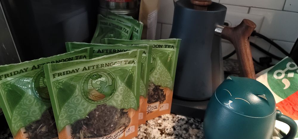 Bunch of bags of Friday Afternoon Tea blends, a Blathers tea towel, green cat steeping mug, and a blue Fellow electric kettle 