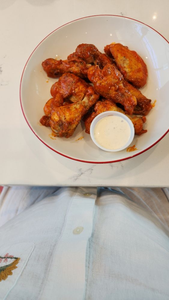 A plate of wings covered in buffalo sauce on a plate. The bottom of the pic shows a clean white shirt.