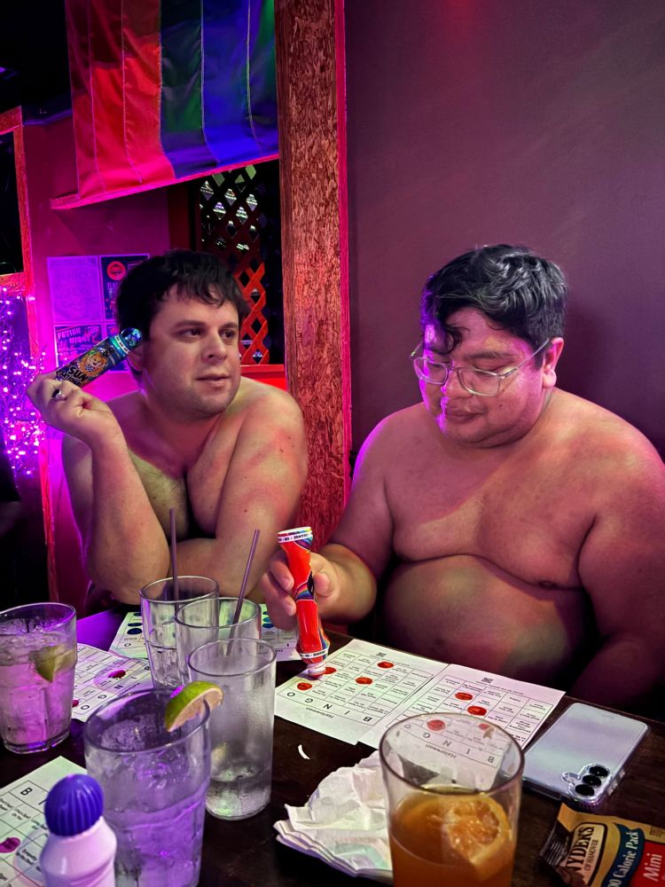 Ricky and I, shirtless at a bar table. On the table are various drinks and bingo cards. Ricky is dobbing his card, while I'm looking off into the distance.