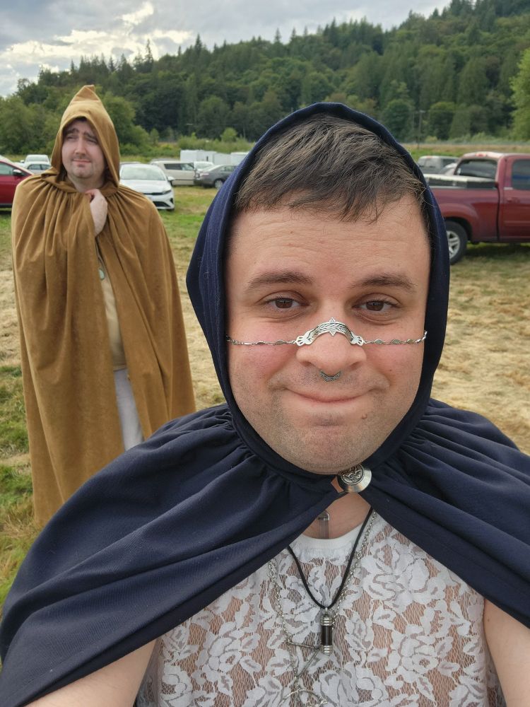Selfie photo of my husband and me. In the front, I'm wearing a white lace shirt and a blue cloak with the hood up. In the back is my husband, wearing a brown hooded cloak.