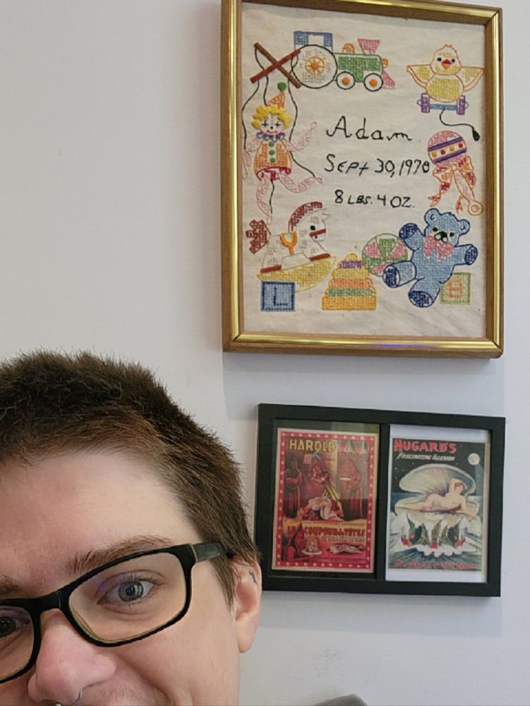 fiidget, a white dude w short hair and classes sits in front of a white wall with some framed art. One is a cross stitch which reads "Adam Sept 30, 197*" its hard to tell if it's supposed to say 1970 or 1978