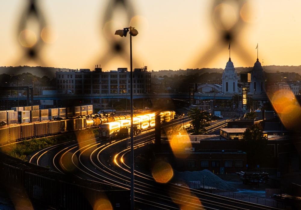A golden hour photo with sun glinting off an MBTA bilevel passenger train in downtown Worcester. The Union Station is in the background, and the CSX intermodal yard is to the left. The trains and station are framed with a blurry shadow of chainlink fence. 