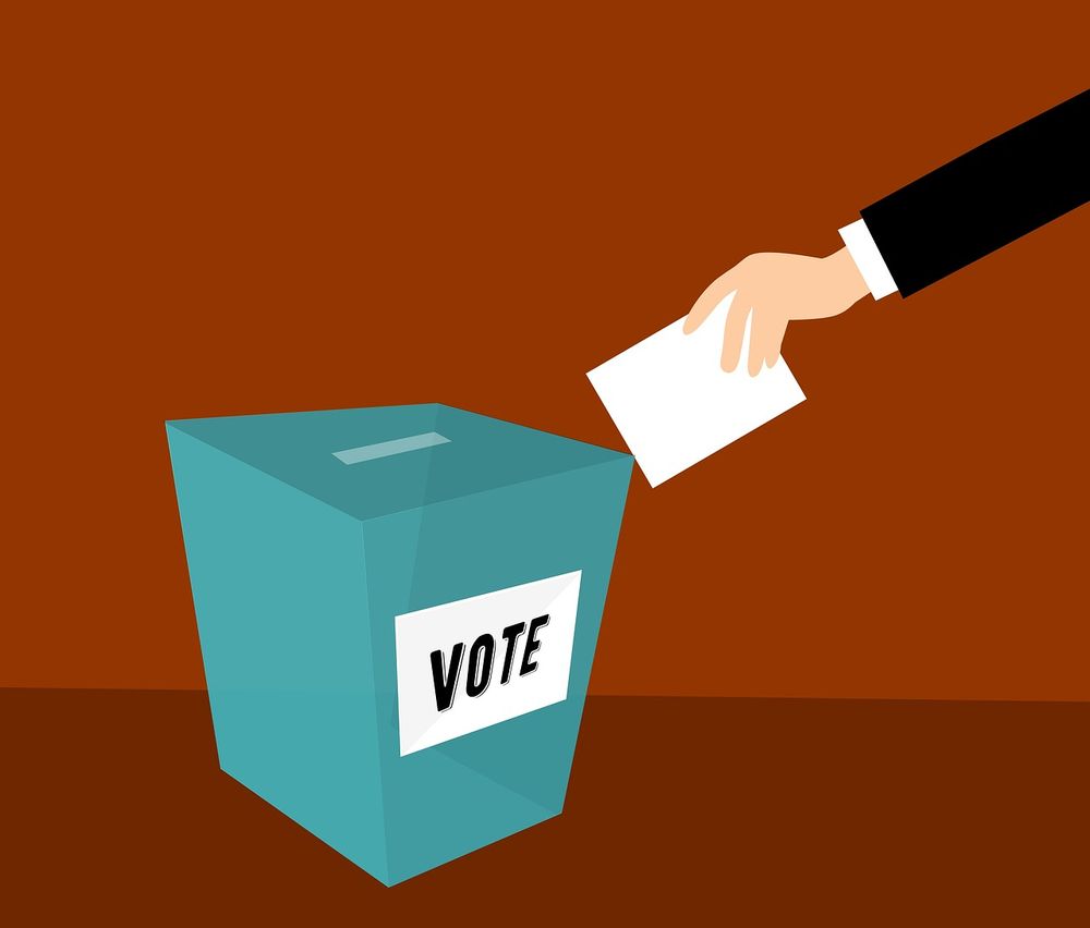 A light-skinned-hand placing a ballot in a ballot box.