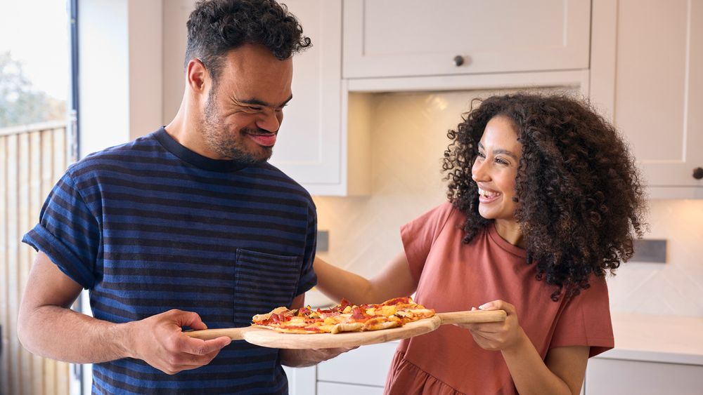 A medium-skinned-man with Down syndrome cooking a pizza with a medium-skinned-woman.