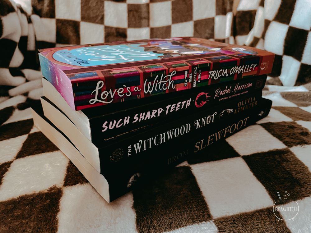 a stack of four books on a checkered blanket: love’s a witch by tricia o’malley, such sharp teeth by rachel harrison, the witchwood knot by olivia atwater and slewfoot: a tale of bewitchery by brom