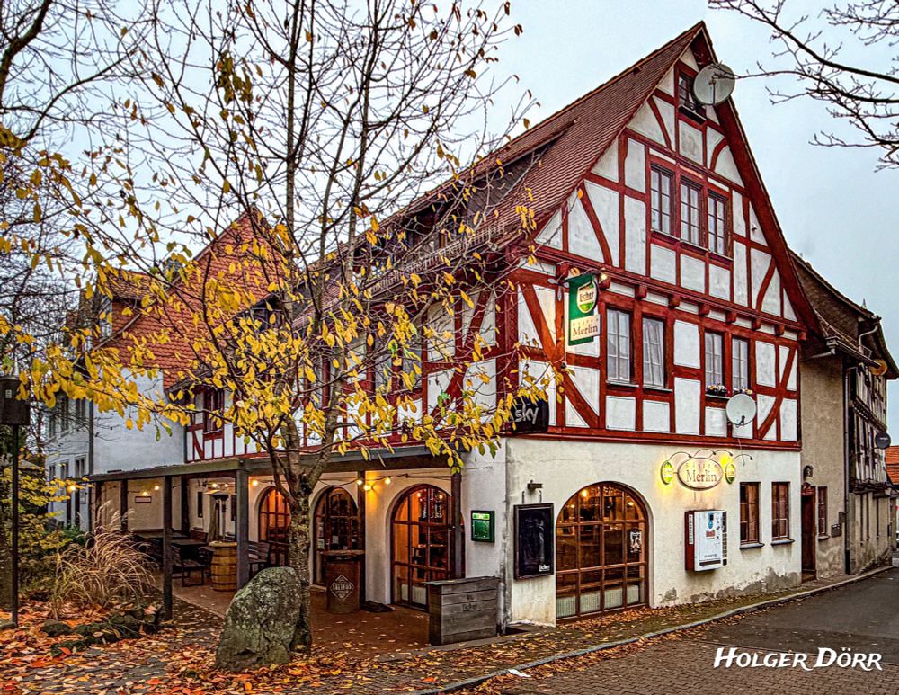 Fachwerkhaus mit rotem Gebälk und weißem Putz in der Altstadt von Grünberg, Hessen. Im Erdgeschoss leuchtet das Bistro Merlin, während herbstliche Blätter auf das nasse Kopfsteinpflaster fallen. Stimmungsvoll beleuchtete Szene an einem kühlen Abend.