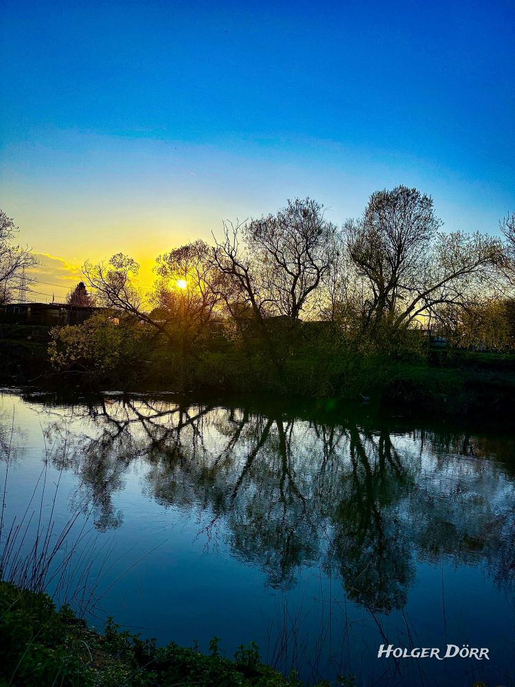 Ein ruhiger Frühlingsabend an der Lahn in Gießen. Die tief stehende Sonne wirft warmes, goldenes Licht durch die kahlen oder gerade austreibenden Äste hoher Bäume. Ihr Licht spiegelt sich sanft auf der glatten Wasseroberfläche des Flusses. Die Szene wirkt still, friedlich und leicht verträumt.