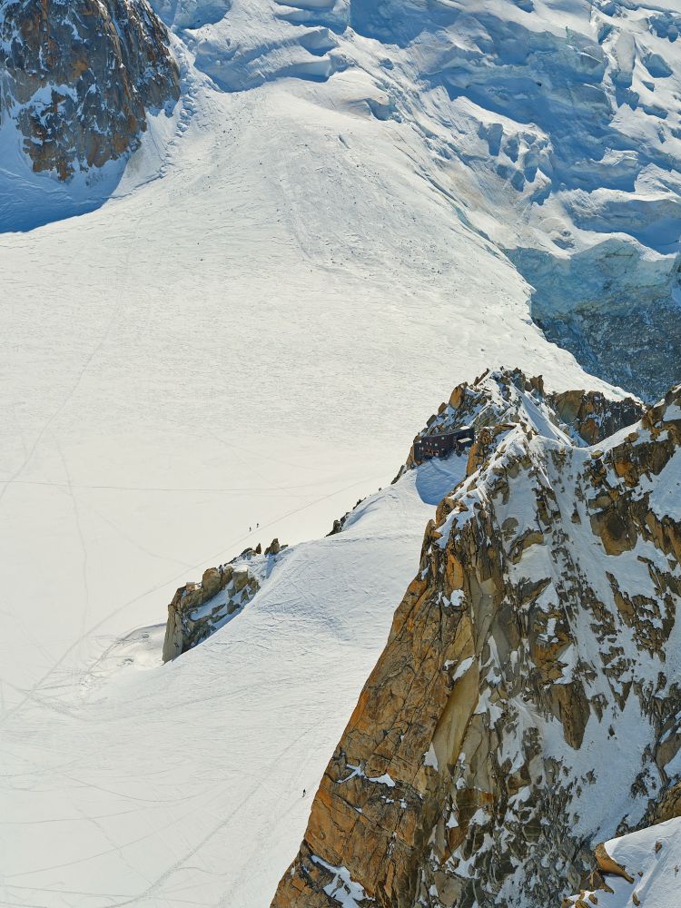 An isometric view of La Refuge Des Cosmiques from L’Aguille Du Midi’s observation deck. Two ski alpinists can be seen traversing the snowfield below the refuge, heading towards Mont Blanc. 