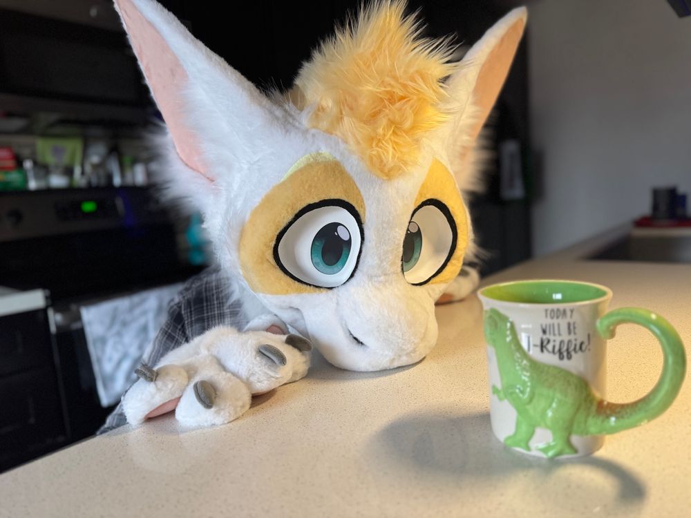 A white and gold furred dragon fursuit leans on a kitchen countertop, staring at a coffee mug. The mug has a T-Rex on it and says “Today will be T-Riffic!”