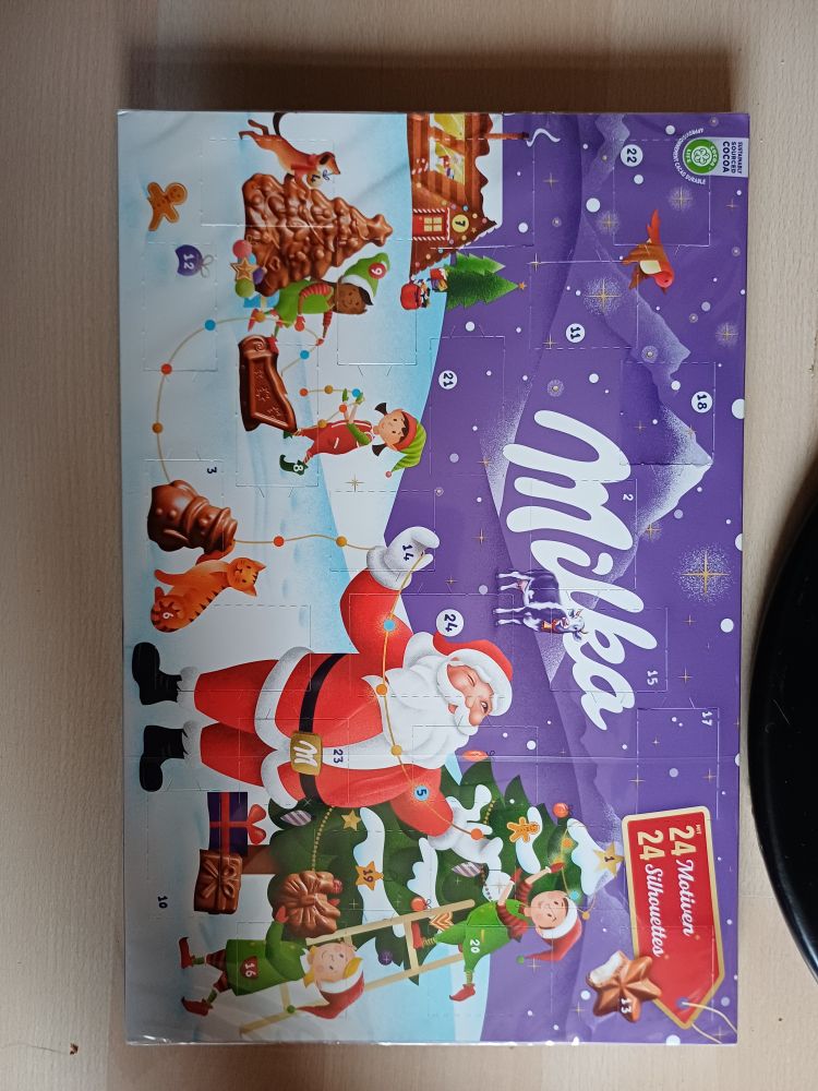 Adventkalender van Milka met 24 vakjes die open kunnen. Op de kalender, die paars is, staat een winters landschap met de kerstman, wat elfjes en een kerstboom.