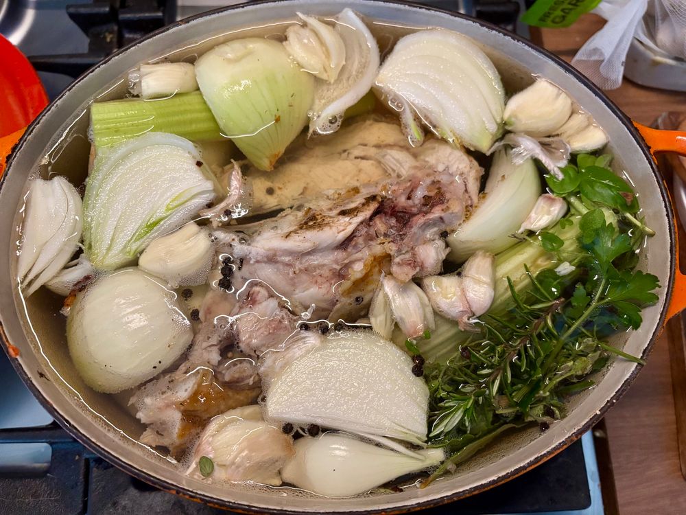 A Dutch oven filled with a chicken carcass, onions, celery, garlic, carrot, herbs, and peppercorns.