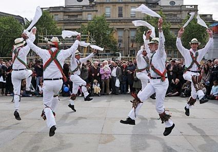 A photo of a nice sunny day in England with some fanciful Morris dancers leaping in the air and flailing their handkerchiefs jubilantly why a crowd watches appreciatively.