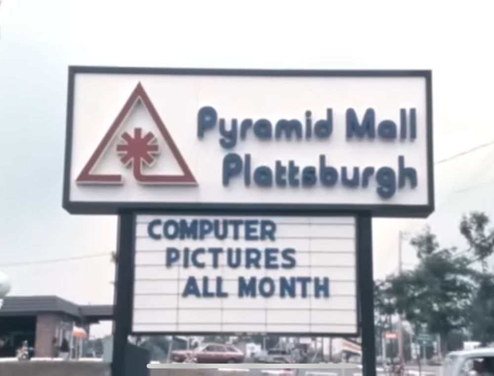 old video still of the sign outside a mall called “Pyramid Mall Plattsburgh” with a logo that is a triangle that has a palm tree or a butthole in it. beneath it another sign reads: COMPUTER PICTURES ALL MONTH