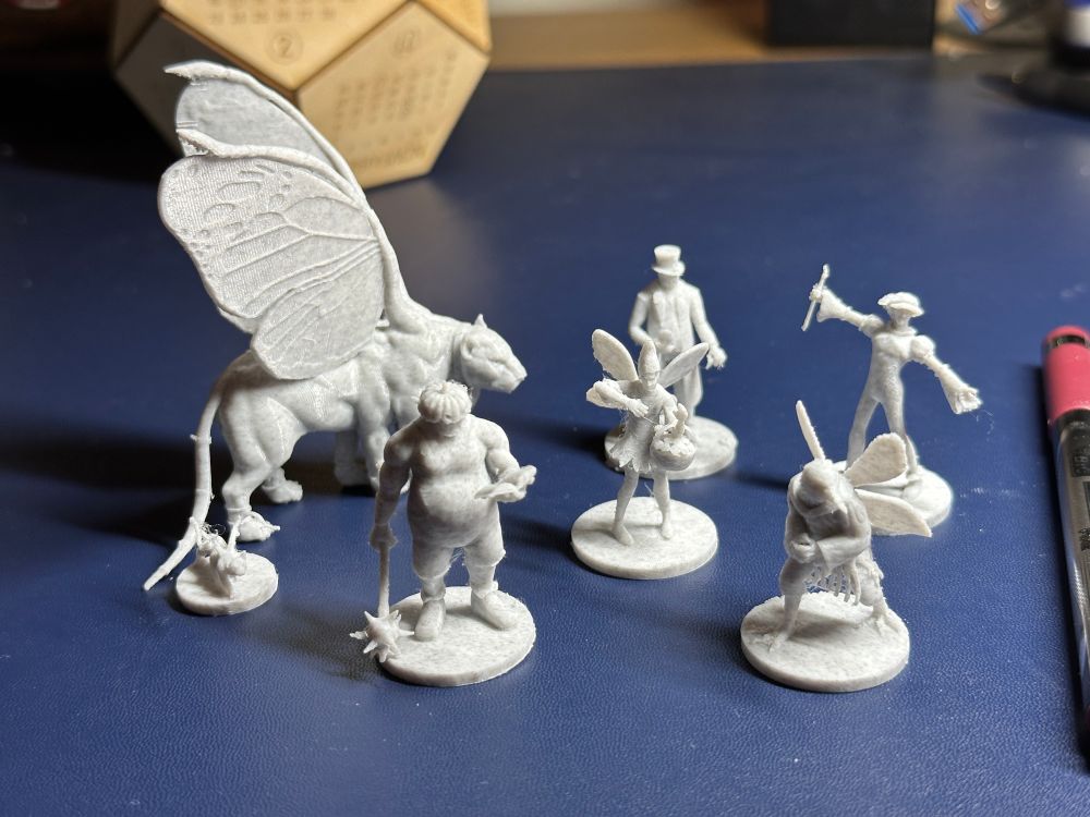 Seven D&D minis on a blue background. The minis were 3D printed with marble PLA filament, and haven’t been cleaned up yet. They are are of the characters from the Wild Beyond the Witchlight D&D adventure module - in particular, the main characters of the Witchlight carnival. 