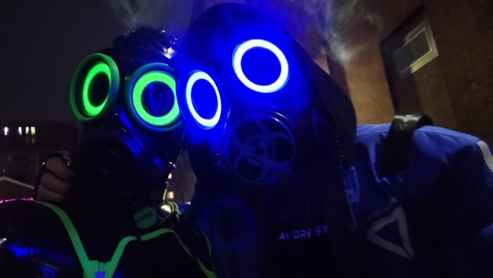 2 drones taking a photo together. One is dressed in latex and is wearing green. The other is wearing blue, has smoke coming out of the mask, and has techwear clothes on top of its amorsey suit.