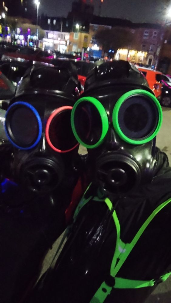 2 people wearing latex and gas masks taking a selfie. The one holding the camera has a primarily green colour scheme on top of their latex, and the other one is wearing red and blue as their colour scheme.