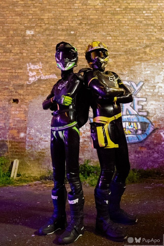 2 people in a mix of rubber and Motocross gear standing back to back with arms folded. The one on the left (JadeDrone) has green accents whilst the one on the right (Natex_xD) has yellow accents.