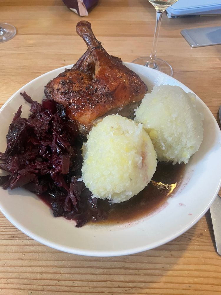 Ein Teller mit gebratener Entenkeule, serviert mit zwei Thüringischer Klößen und einer Beilage Rotkohl. Ein Glas Weißwein ist im Hintergrund auf einem Holztisch zu sehen.