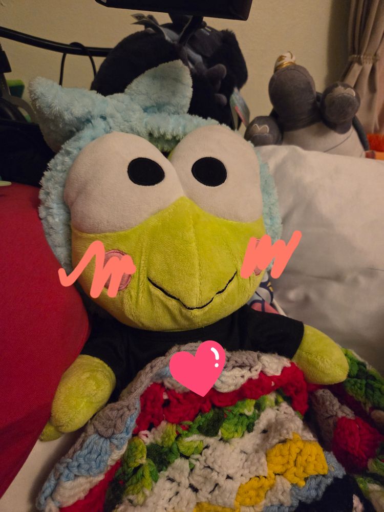 a stuffed keroppi plush wearing a blue headband and tucked under a multicolored blanket. a heart emoji and pink squiggles on his cheeks for blush have been added