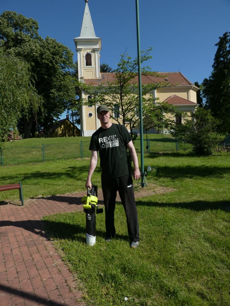 Image of Project leader, Péter Batáry, HUN-REN Centre for Ecological Research working with an insect suction sampler in the village centre of Salköveskút, near the city Szombathely, Hungary. He looks like he is on a lush, green, grassy area with tall trees and a church in the background. Attila Torma