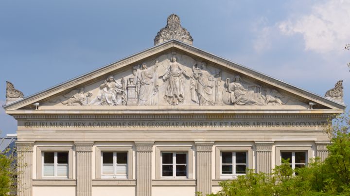 Giebelrelief unseres Aulagebäudes am Wilhelmsplatz (Copyright: Frank Stefan Kimmel)

Dargestellt ist im Detail: Im Zentrum schwebt der geflügelte Genius der Wissenschaft. Die vier Figuren zu beiden Seiten symbolisieren die klassischen Fakultäten der Universität: links außen die Medizin mit Schale, Schlange und der Fackel des Lebens; daneben die Theologie mit Kreuz, Kelch und den Gebotetafeln; rechts vom Genius die Jurisprudenz mit Waage, Gesetzbuch und Richtschwert; rechts außen auf die Weltkugel gestützt die Philosophie mit einer Leier in der Hand. Eine Sphinx mit Totenschädel, Sinnbild für die Rätsel der Natur, sowie ein Greif, Symbol der Fantasie, schließen die Darstellung nach außen hin ab. 