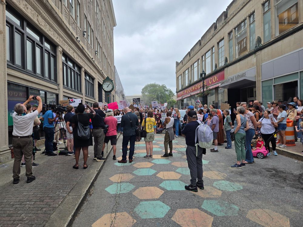 People gathered in a street around a speaker at a press conference, taking photos and holding signs of protest. In the background is a building with an old-fashioned sign proclaiming F. W. Woolworth Co. in gold letters.