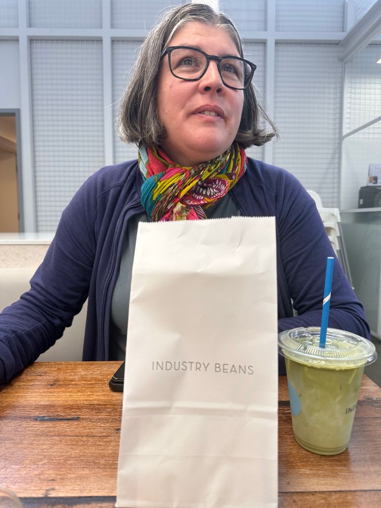 The same woman looking wistfully into the distance waiting for a cookie to be delivered, sat at a bench table with a white bag lndustry Beans in front of her