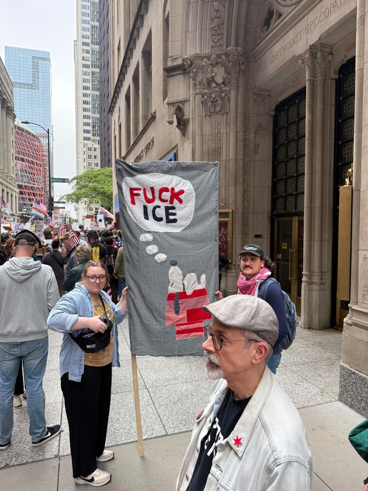 Photo of a a street full of protesters. A quilted sign shows Snoopy sleeping on his dog house with a thought bubble that says “Fuck ICE”