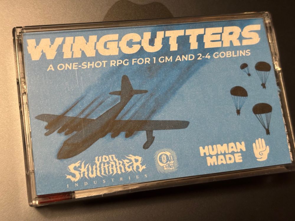 A cassette tape case containing a game titled Wingcutters: a one-shot RPG for 1 GM and 2-4 goblins.  It features a blue background with a crashing plane and 4 goblins parachuting in the far distance.