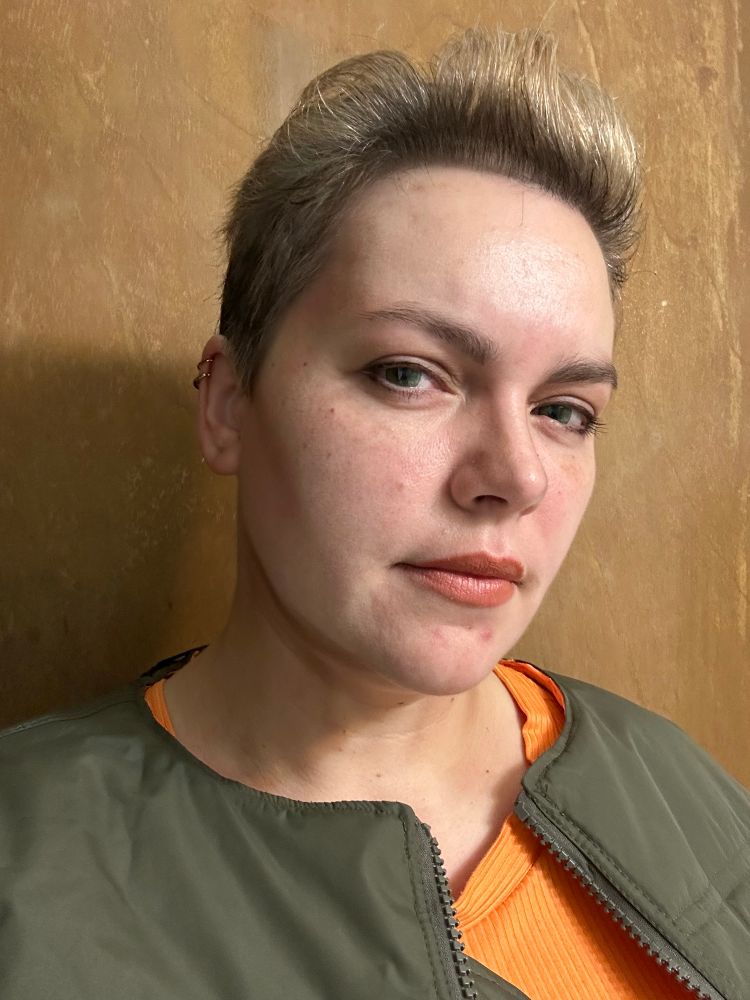 A selfie of me dressed as Korben Dallas from The Fifth Element. I have an orange tank top and my modified olive green flight jacket on. My hair is dyed blonde and I have green contacts on, giving the camera a bit of a stink eye