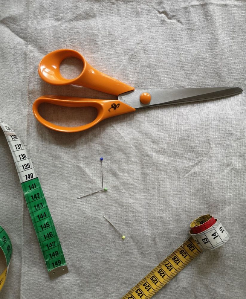 Kuvassa värjäämätöntä pellavakangasta, mittanauha, nuppineuloja ja sakset.