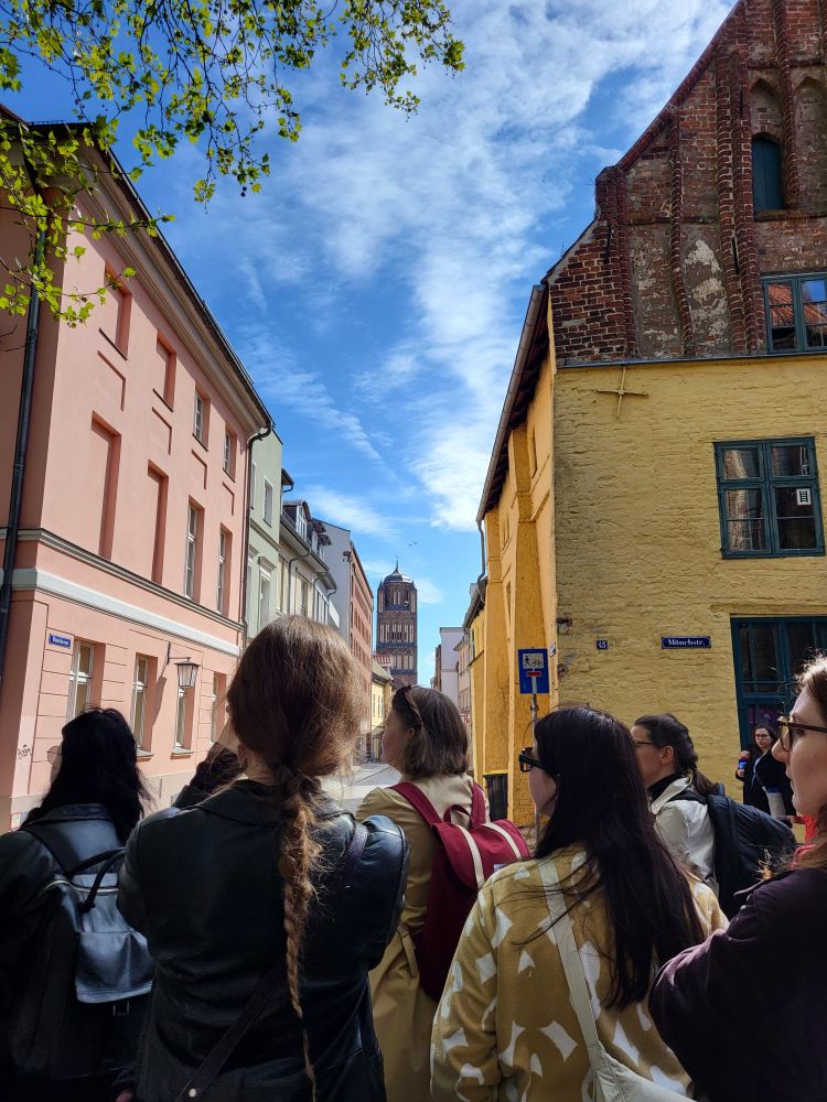 Group of people in front of an old town street in Stralsund. Left an 18th-century pink house, right a medieval brick house with yellow paint, in the background the gothic tower of St. James