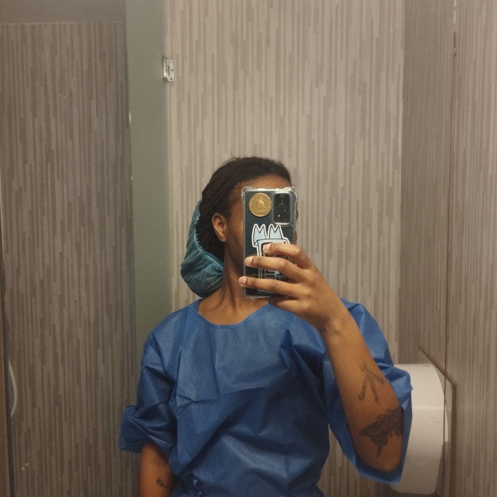 Mirror selfie en robe d'hôpital bleue foncée. Mon visage est caché par mon téléphone où dans la coque il y a ma médaille des 18 mois. La charlotte tient mes cheveux en un bas chignons mais le reste de la tête n'est pas couvert 