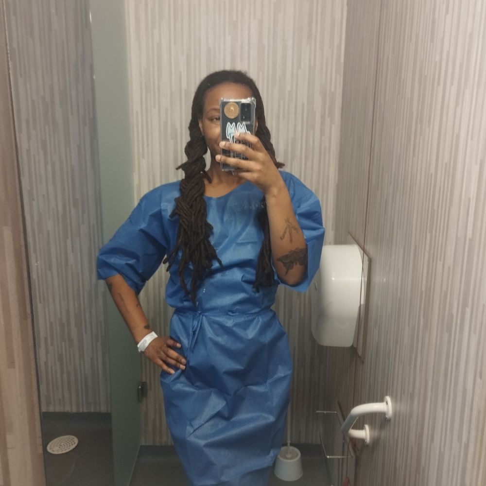 Mirror selfie en blouse bleue d'hôpital. Une main sur la hanche, l'autre qui tient mon téléphone qui cache une partie de mon visage. J'ai deux longues tresses de chaque côté 
