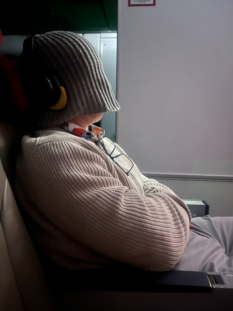 A man sleeping on an airplane. He's wearing a thick knitted sweater and has a ribbed, knitted beanie pulled down to cover his face with headphones outside the beanie.