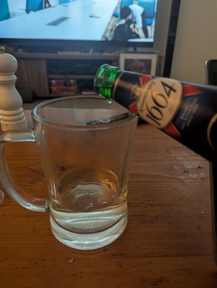 A pint glass with an inch of clear liquid (soju) in it, with a bottle of Kronenbourg beer tilted against the rim, about to be poured in