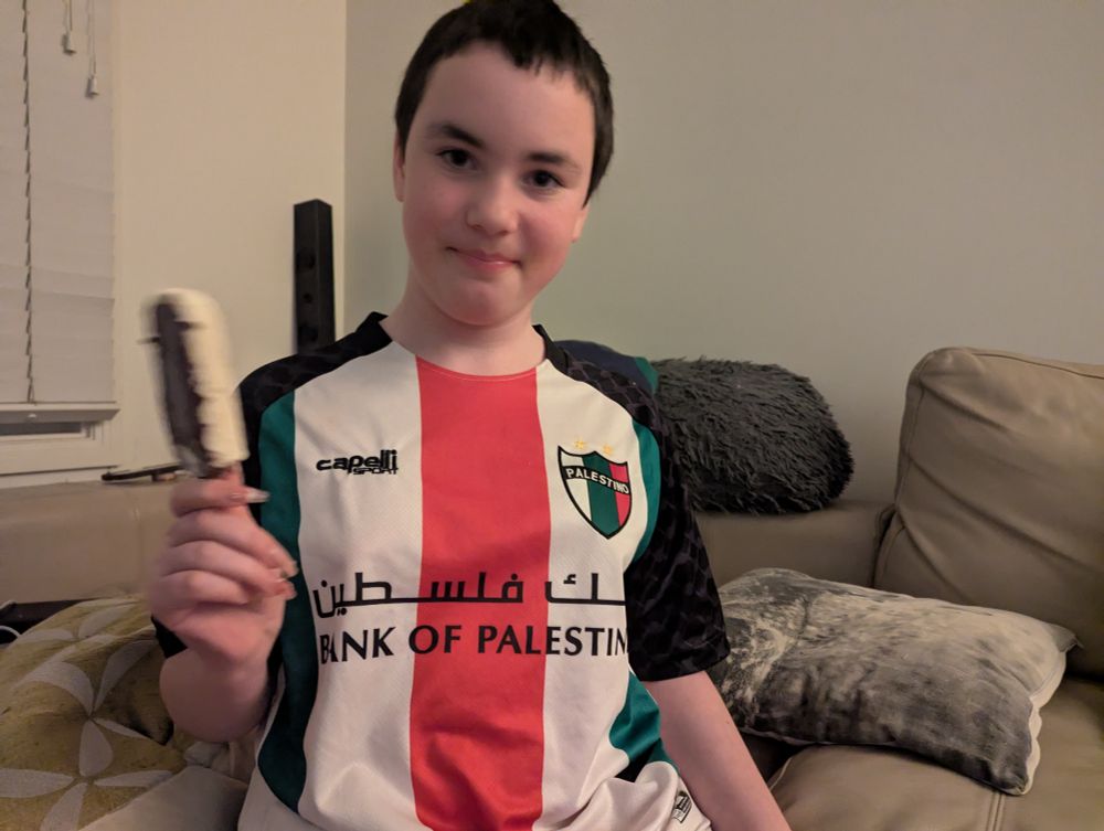 A smiling, gorgeous crew cut 10yo enby eating an ice cream, wearing a Palestino football jersey with "Bank of Palestine" emblazoned on it in English and Arabic. 