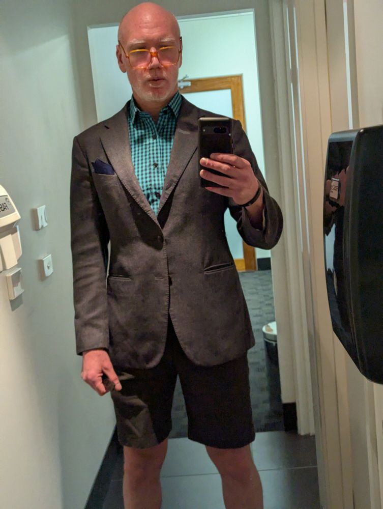 A bald man in a mirror selfie, wearing a grey suit jacket and matching tailored shorts with a blue check shirt.
